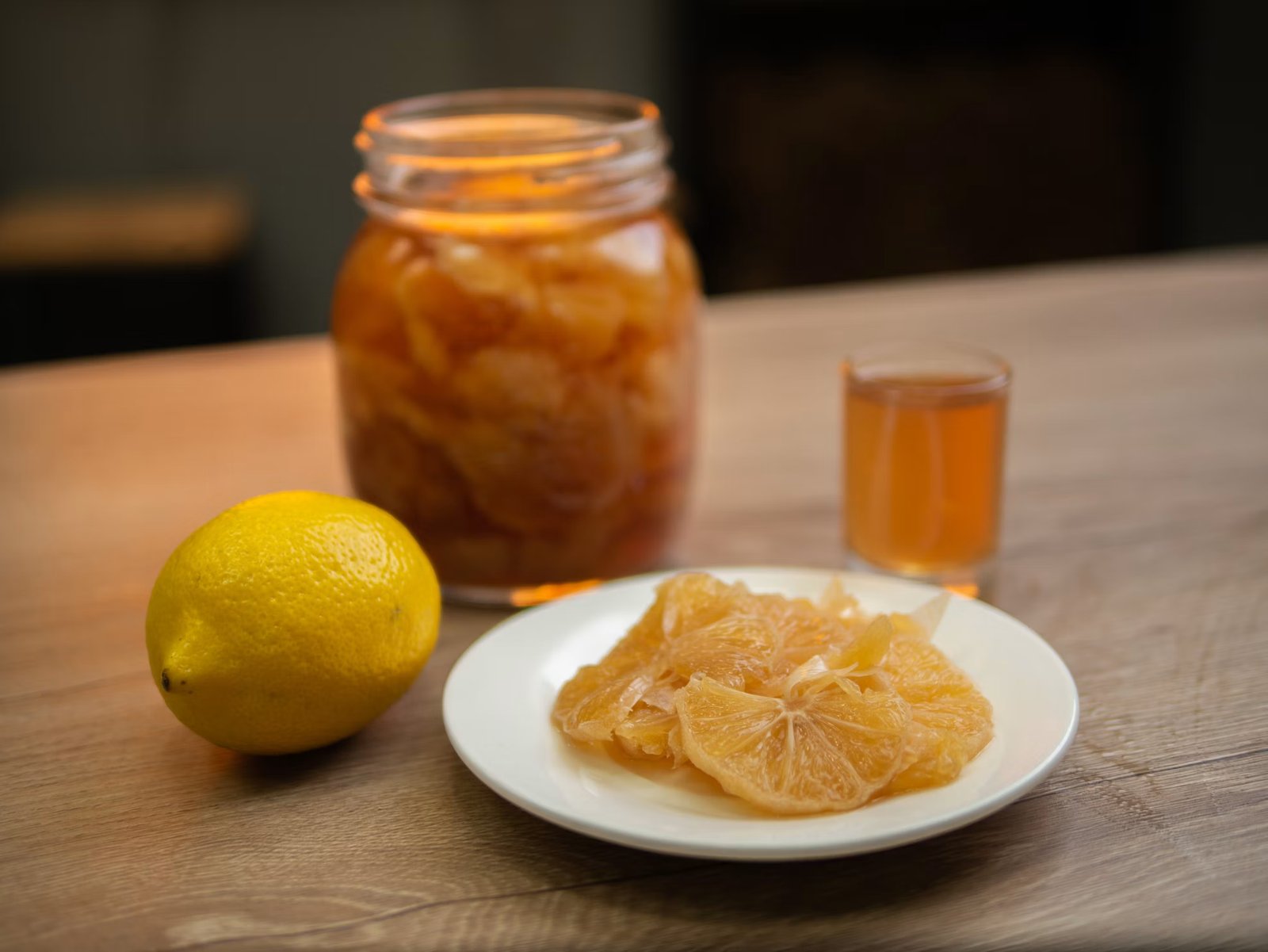 Jar of sweet lemon pickle in jaggery syrup beside a fresh lemon, a small glass of the golden syrup and a plate of jaggery-cured lemon slices